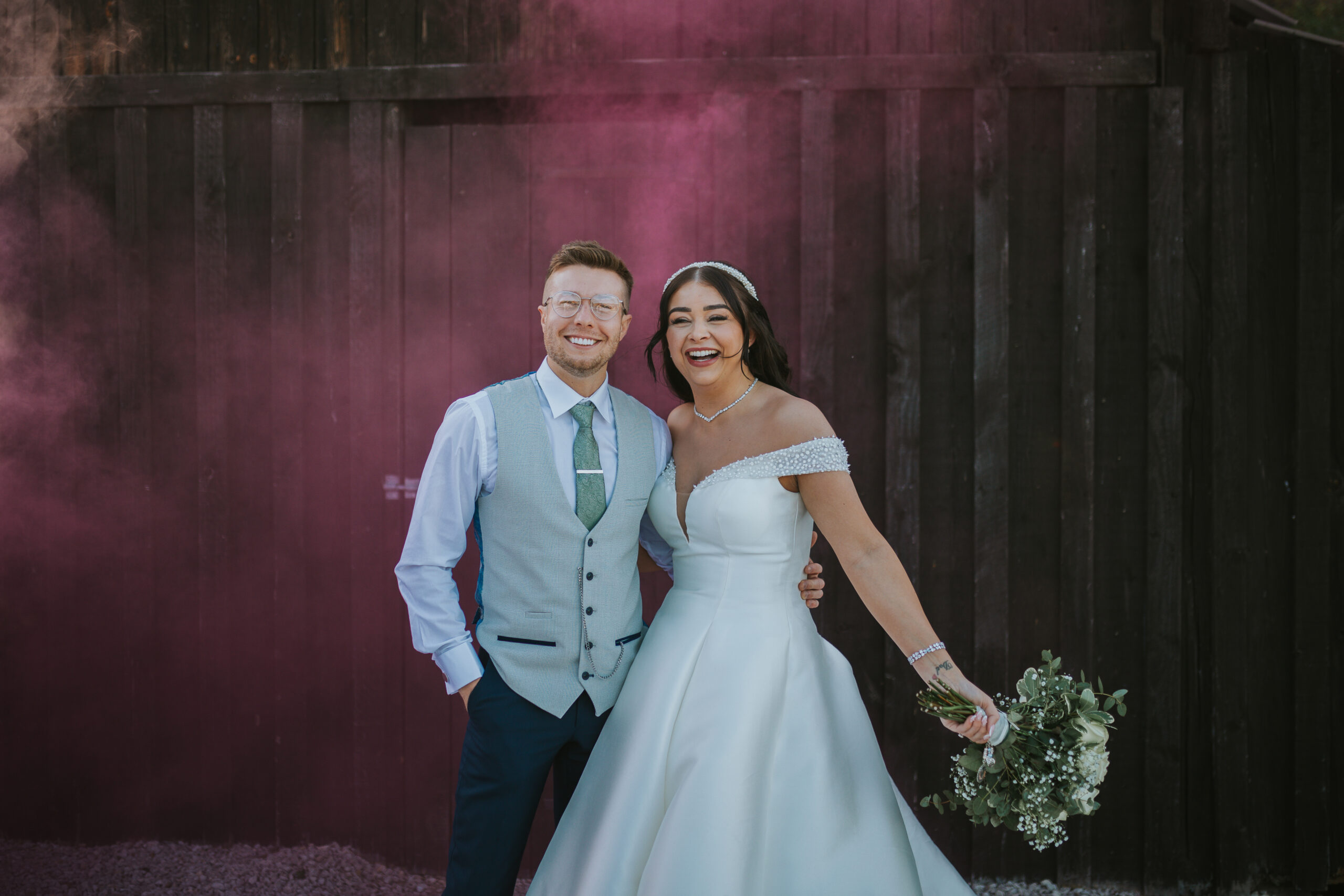 Recently married couple smiling. The bride holding a bouquet with a smoke bomb in the background. Photographed at Hazel Gap Barn, by Slate Cottage Studio.