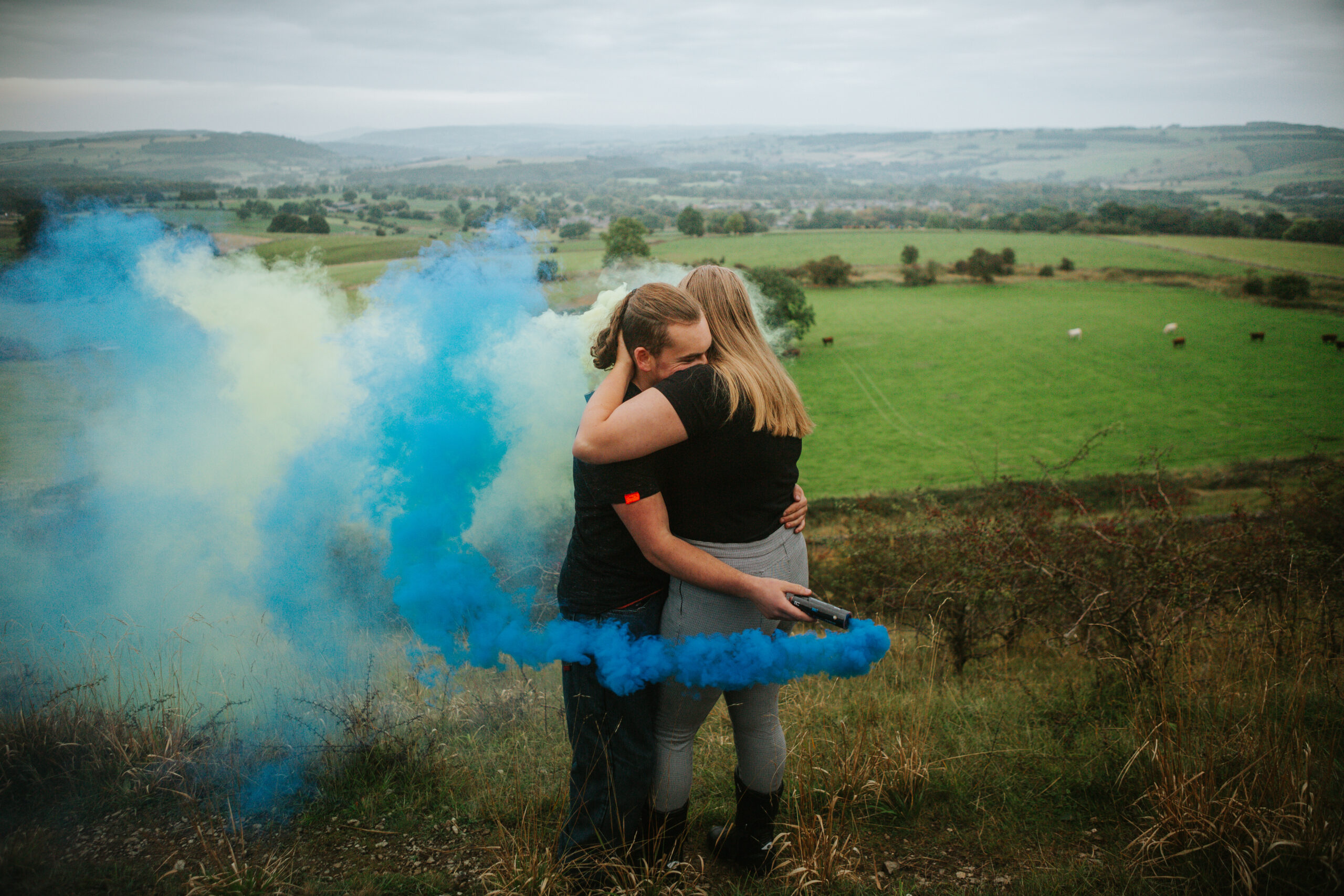 Couple hugging surrounded by blue smoke during a Peak District surprise proposal, adding a creative and dramatic flair to the engagement photography session.