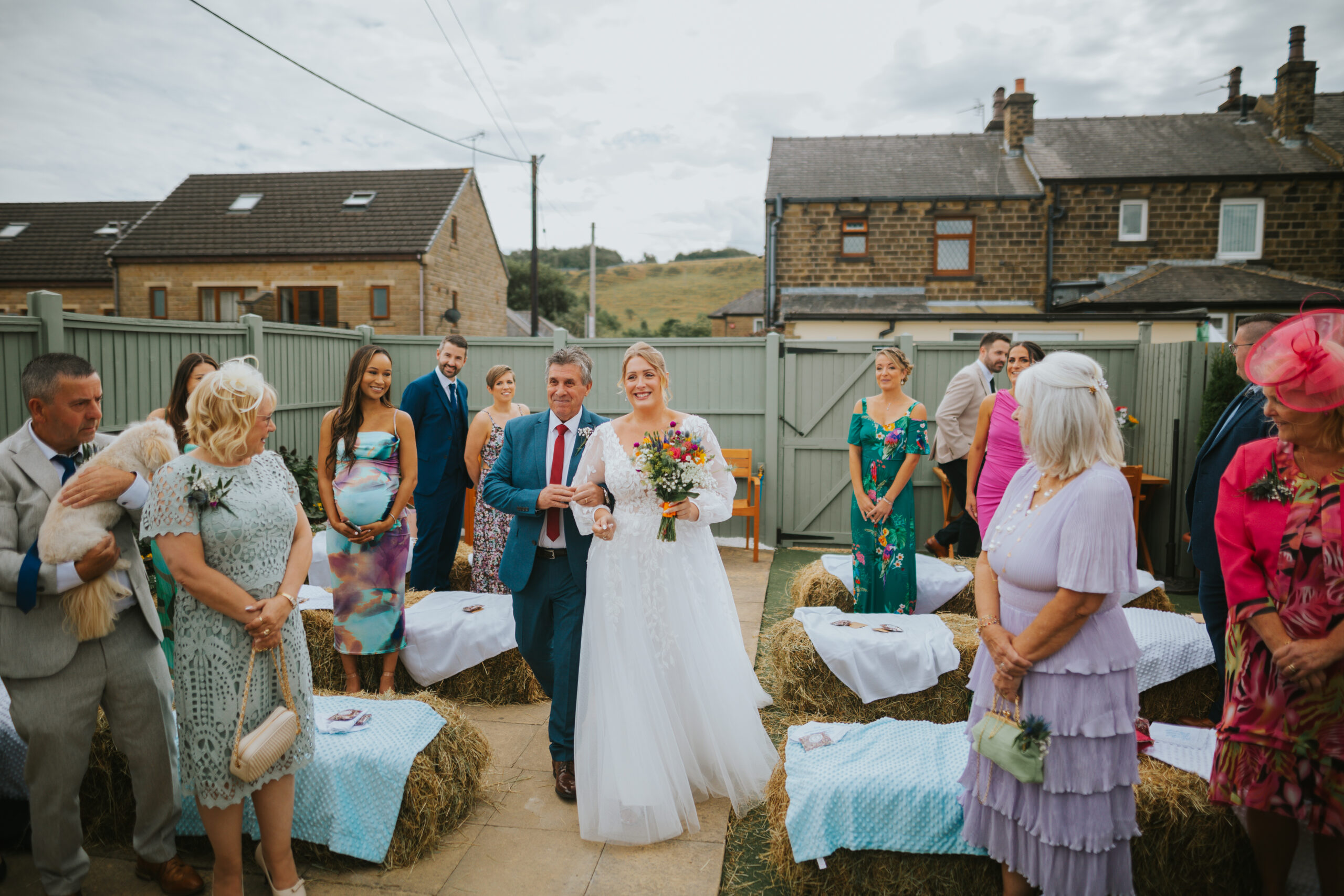 Bride walking down the aisle with her father at an intimate back garden wedding, surrounded by hay bale seating and smiling guests, capturing relaxed and alternative wedding vibes.