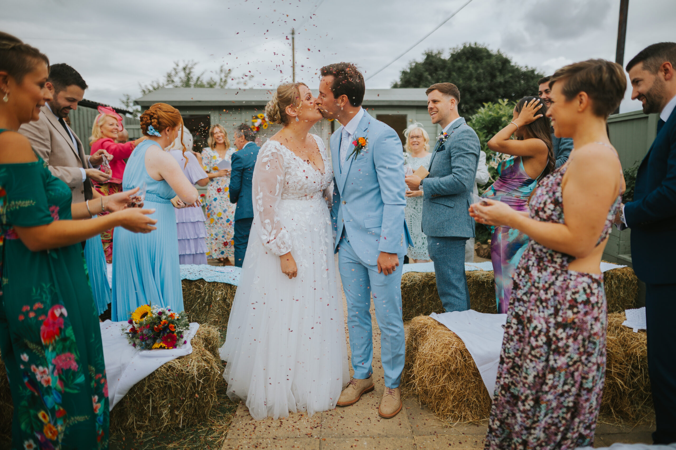 Newlywed couple sharing their first kiss during a back garden wedding ceremony, surrounded by friends and family celebrating with confetti—a beautiful moment in an alternative outdoor setting.