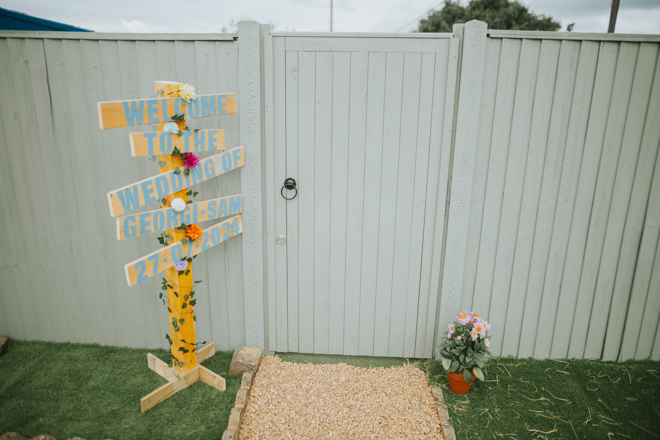 Hand-painted welcome sign at a back garden wedding featuring colourful florals, creating a unique and personal touch for a DIY celebration. Perfect for alternative wedding ideas.