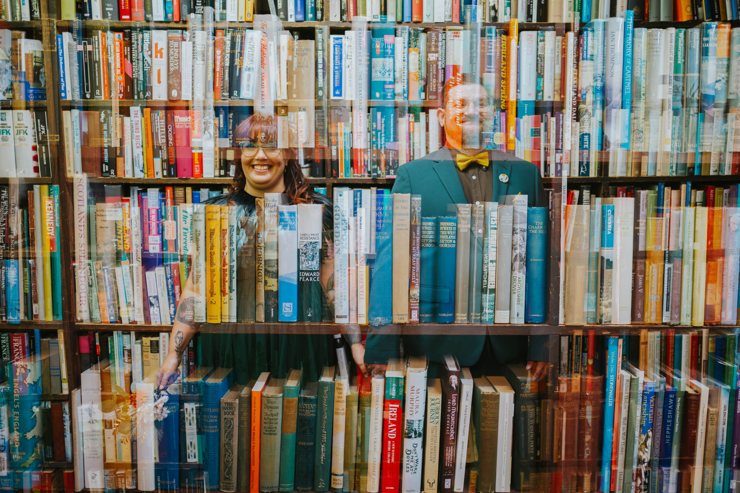 Creative double-exposure photo of a couple smiling behind colorful shelves of books, showcasing their love for unique and alternative wedding photography.