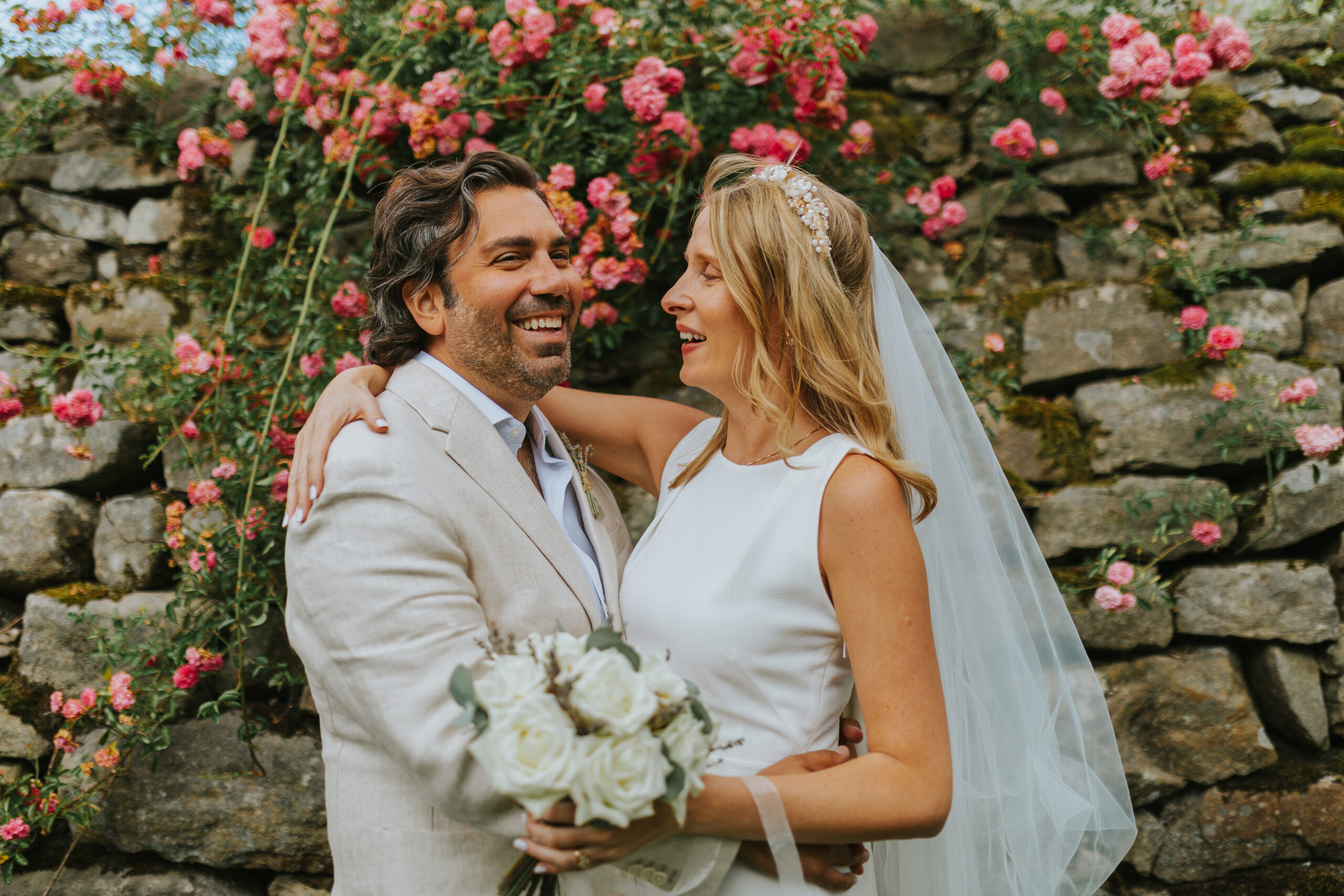 Bride and groom sharing a romantic moment at The Priests House Skipton, surrounded by vibrant pink roses and a historic stone wall in the Yorkshire Dales. Perfect setting for natural, relaxed wedding photography. Photographed by Slate Cottage Studio.