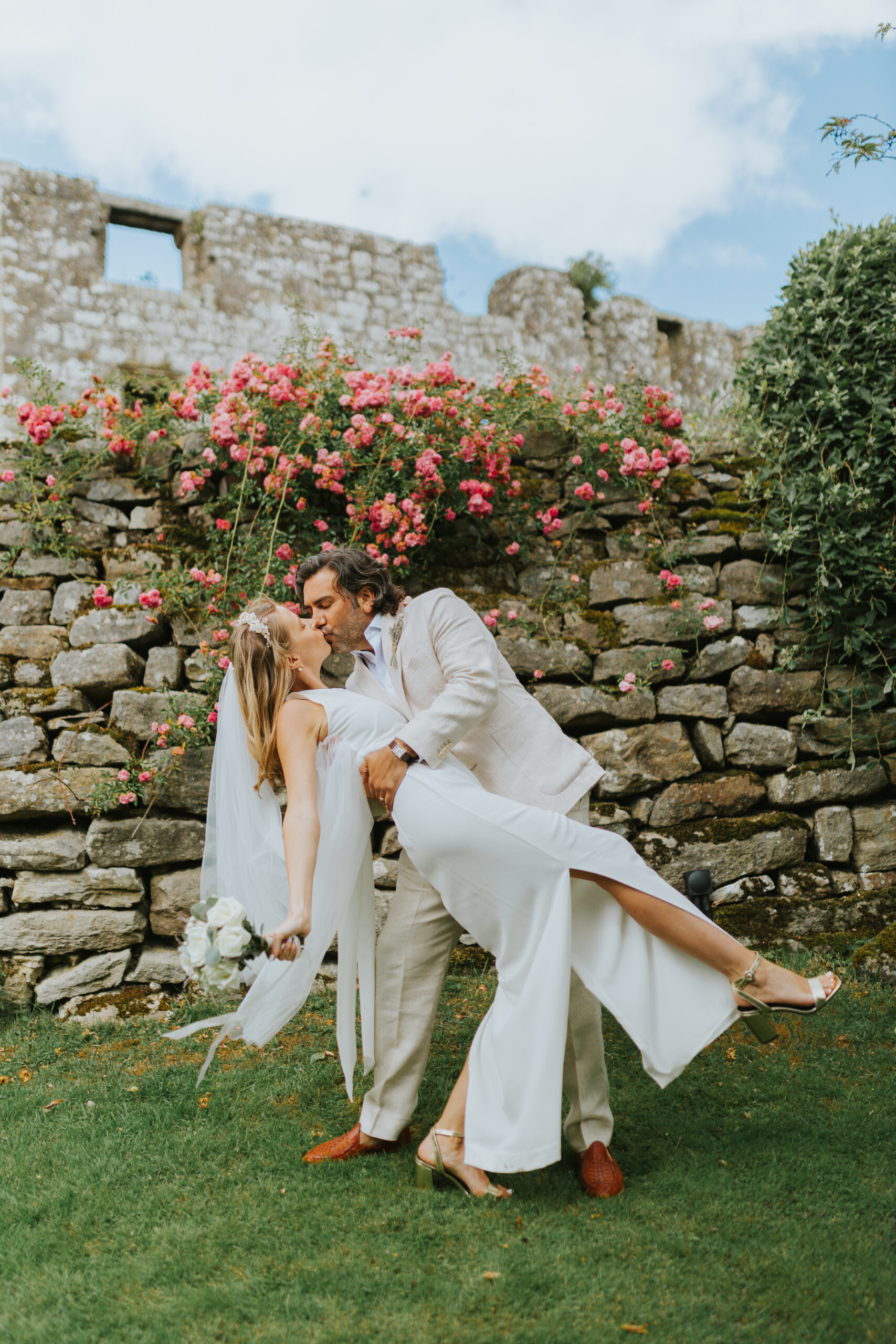 Bride and groom sharing a romantic kiss at The Priests House Skipton, surrounded by vibrant pink roses and a historic stone wall in the Yorkshire Dales. Perfect setting for natural, relaxed wedding photography. Photographed by Slate Cottage Studio.